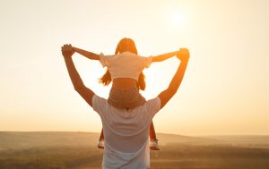 Back view of a father carrying his daughter on neck and enjoying freedom while spending time together in nature against sunset sky